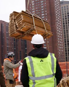 Hillmann Consulting project manager overseeing construction site operations as workers coordinate the safe lifting of building materials in an urban high-rise environment.