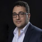 Professional headshot of John Lauretta wearing glasses, a navy blazer, and a light-patterned dress shirt, set against a dark background.