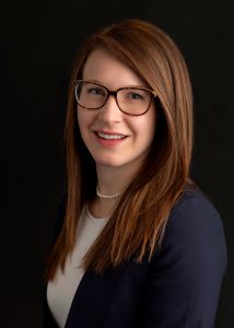 Professional headshot of Kristen Brink, PE, Principal/Partner at Hillmann Consulting, wearing glasses and a navy blazer against a dark background.