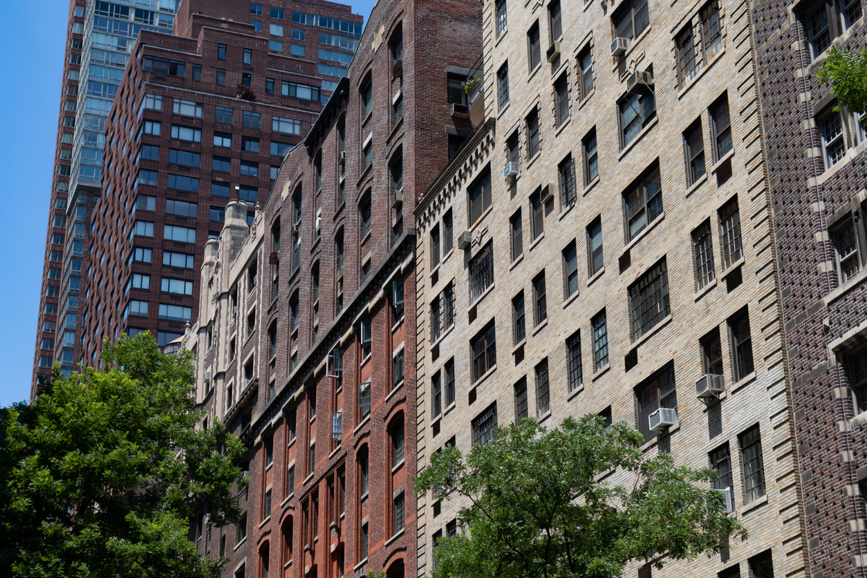 Historic brick apartment buildings with window air conditioners in a dense urban neighborhood, surrounded by trees and high-rise towers in the background on a sunny day.