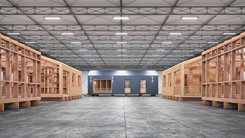 Modular homes being constructed inside a large factory warehouse, showing multiple wood-framed units in production alongside a finished modular structure, representing the efficiency and precision of modern modular construction.