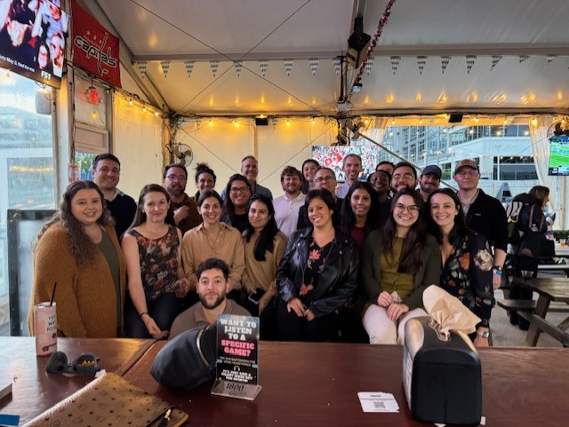 Large group of smiling Hillmann colleagues gathered around a long table at an outdoor restaurant or beer garden, posing for a group photo during a team social event under string lights.