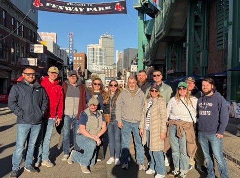 Hillmann team members posing outside Fenway Park on a sunny day in Boston, standing beneath the iconic stadium entrance sign during a group outing.