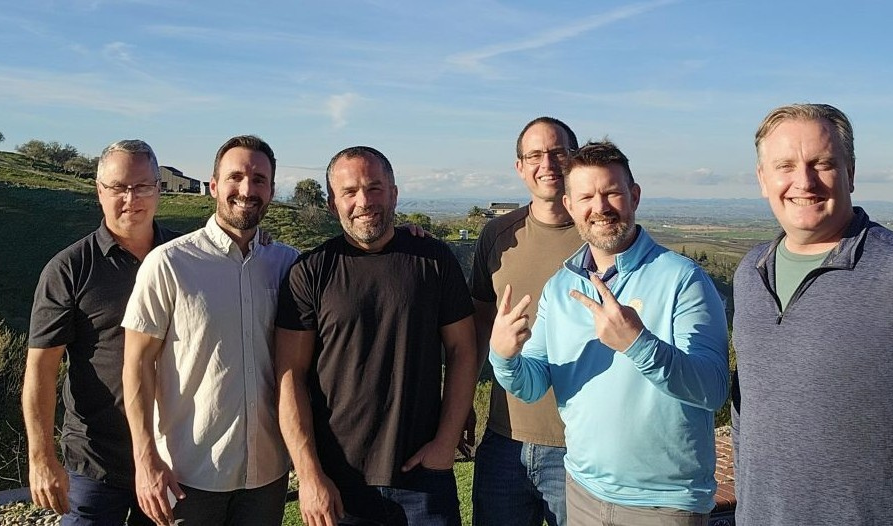 Team members from Hillmann Energy Consulting posing together outdoors on a sunny day with scenic hillside and valley views in the background during a company gathering.