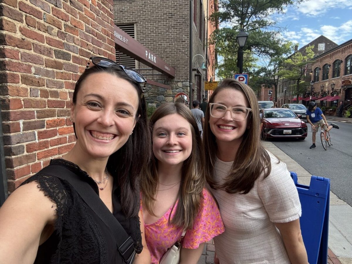 Three Hillmann employees smiling for a selfie outdoors along a brick sidewalk, with parked cars and residential buildings visible in the background on a sunny day.